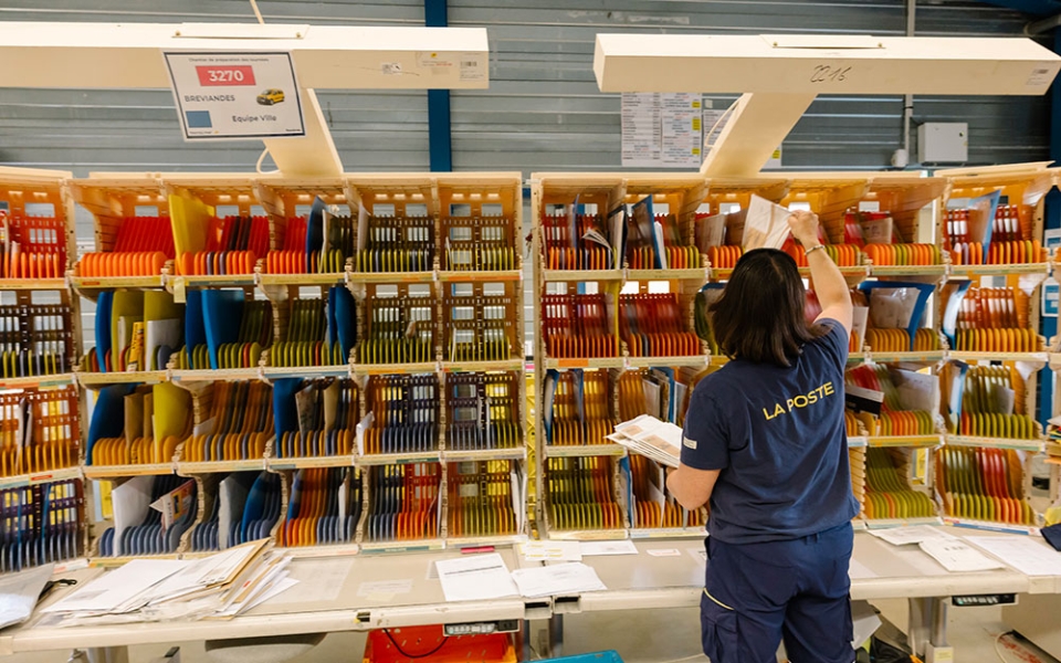 Un facteur ou un « collègue préparateur » commence la tournée… devant des casiers (chaque case correspond à un « point de distribution »).